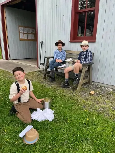 Three boys in hats and suspenders eating lunch from tin pails outside a white building