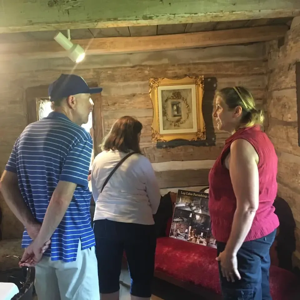 A guide chatting with two visitors inside the log cabin, a gilt-framed portrait on the wall