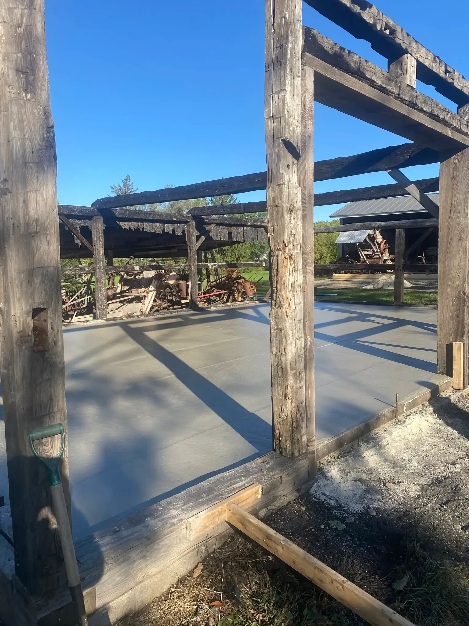 The finished concrete floor curing inside the shed's standing charred frame, a shovel leaning against a post and new roof trusses staged in the background