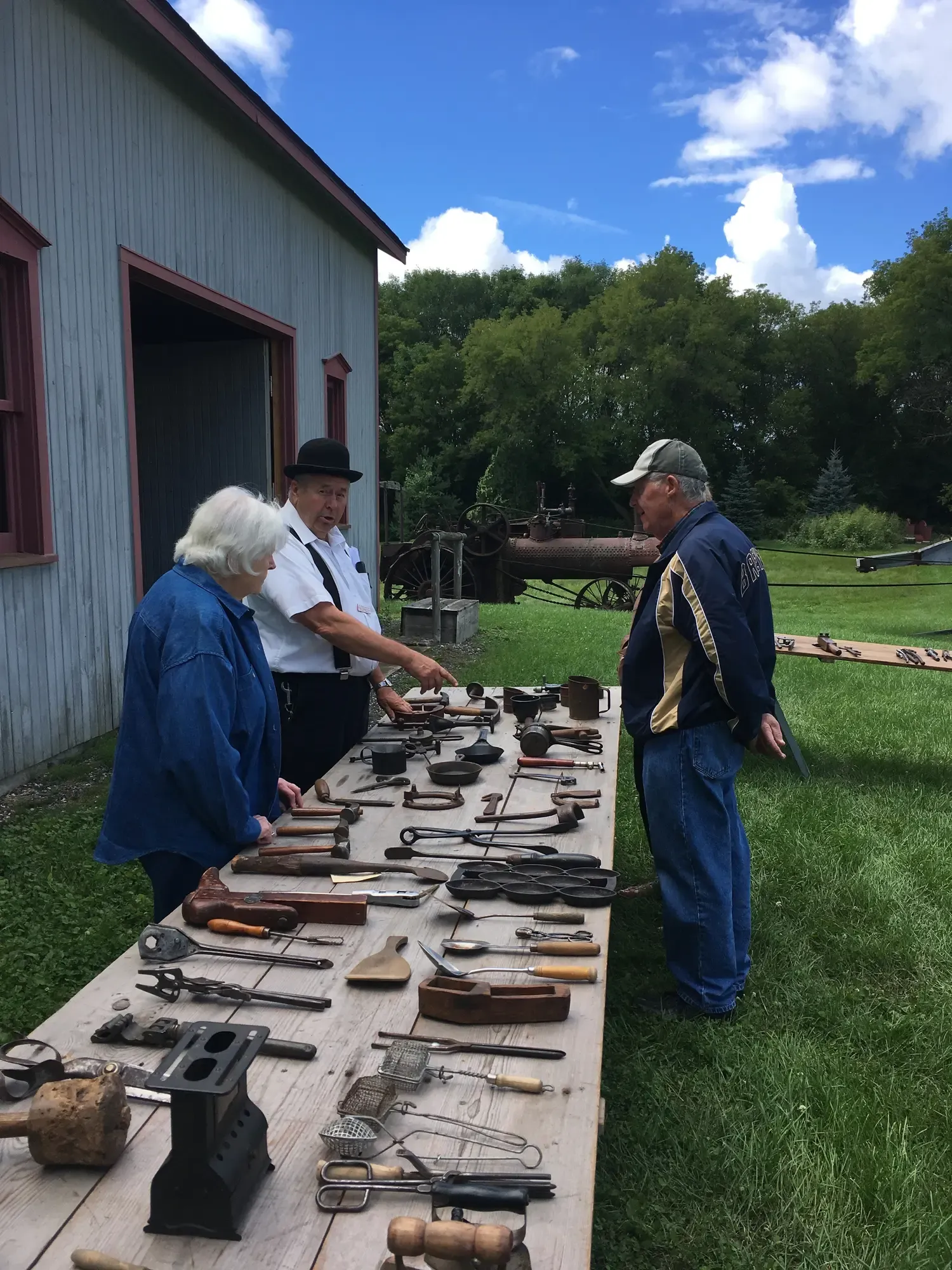 A society member in bowler hat and suspenders walking visitors through a table of antique kitchen tools