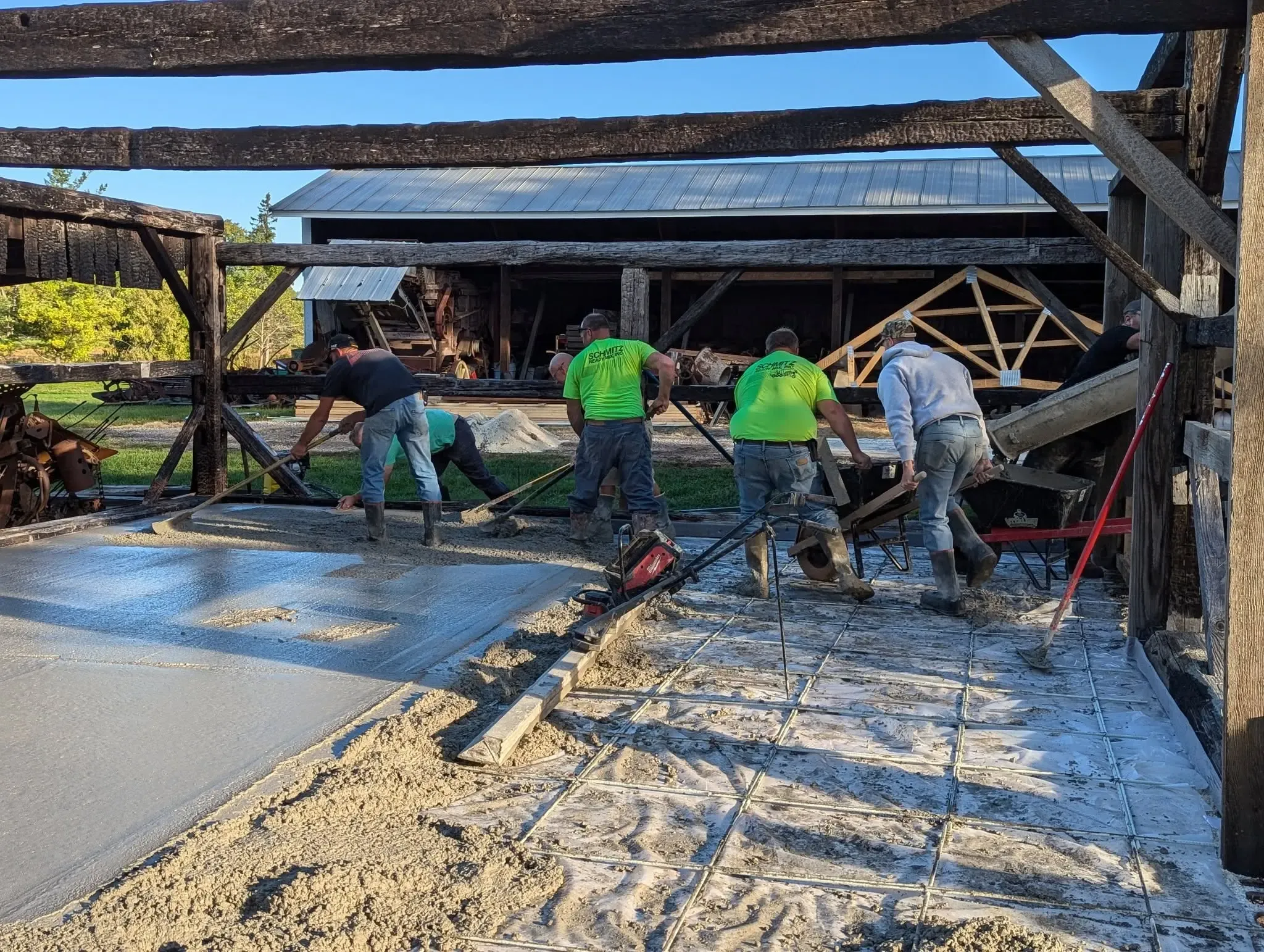 Volunteers and a concrete crew screeding a fresh concrete floor inside the Mueller Shed's charred timber frame, wheelbarrows carrying concrete in from a mixer chute