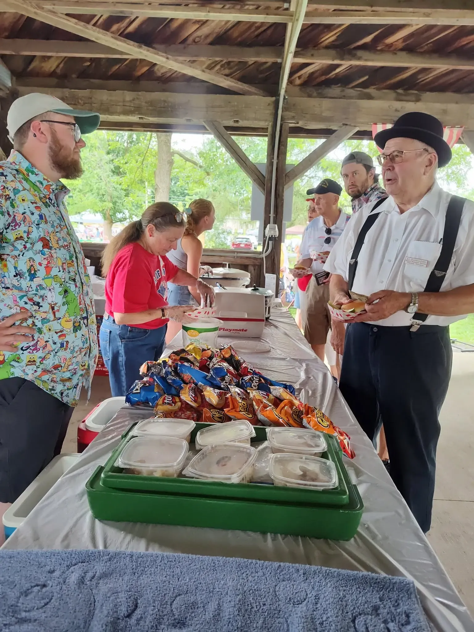 Volunteers dishing up plates at the picnic shelter, a member in bowler hat and suspenders first in line