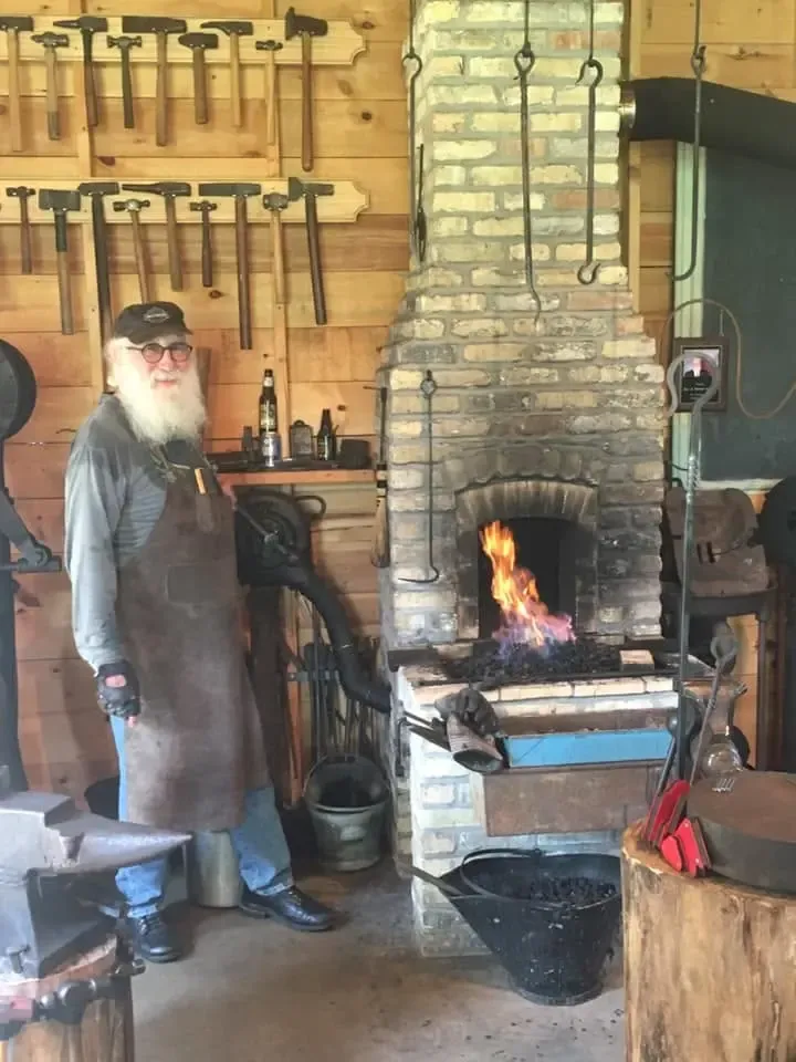The blacksmith in his leather apron beside the brick forge, flames rising from the firepot, with rows of hammers and tongs hung on the wall