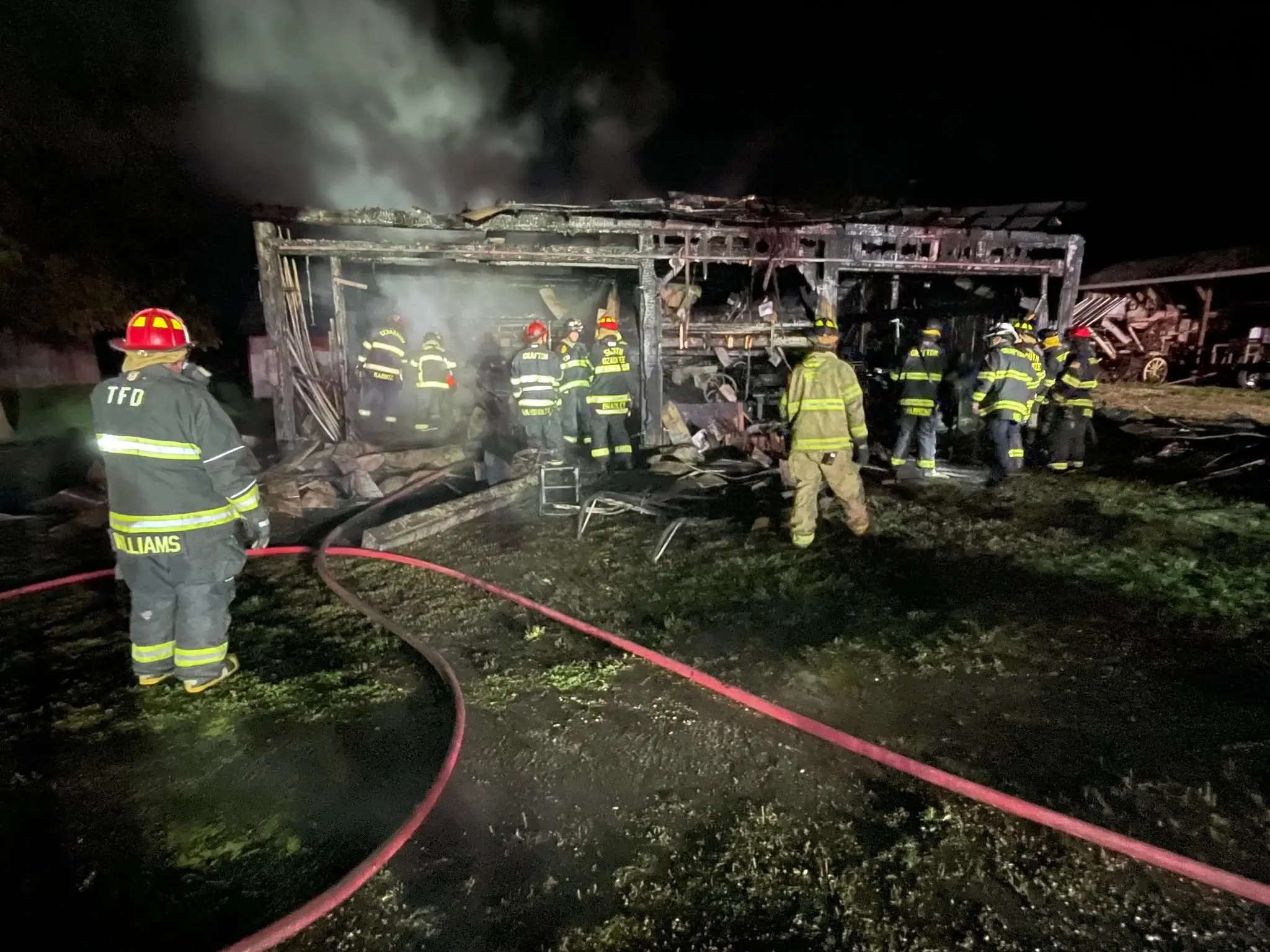 Firefighters from several departments working through the charred frame of the Mueller Shed in the dark, hose lines crossing the wet grass and smoke still rising from the roofline