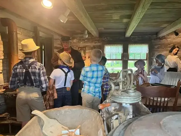 A docent talking with schoolchildren inside one of the historic buildings, antique tools all around