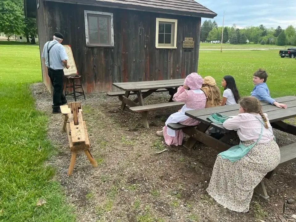 A schoolmaster teaching from a chart outside a weathered shed while children in pioneer dress look on from picnic tables