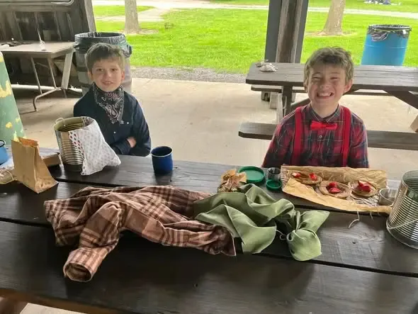 Two boys in pioneer costume grinning over cloth-wrapped lunches at a picnic table