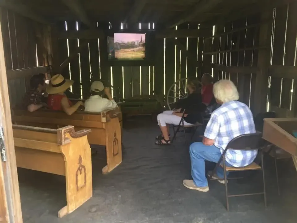 Visitors seated on wooden pews inside the rustic board-walled shed, watching a film about the historic site on a screen, a wagon wheel leaning against the back wall