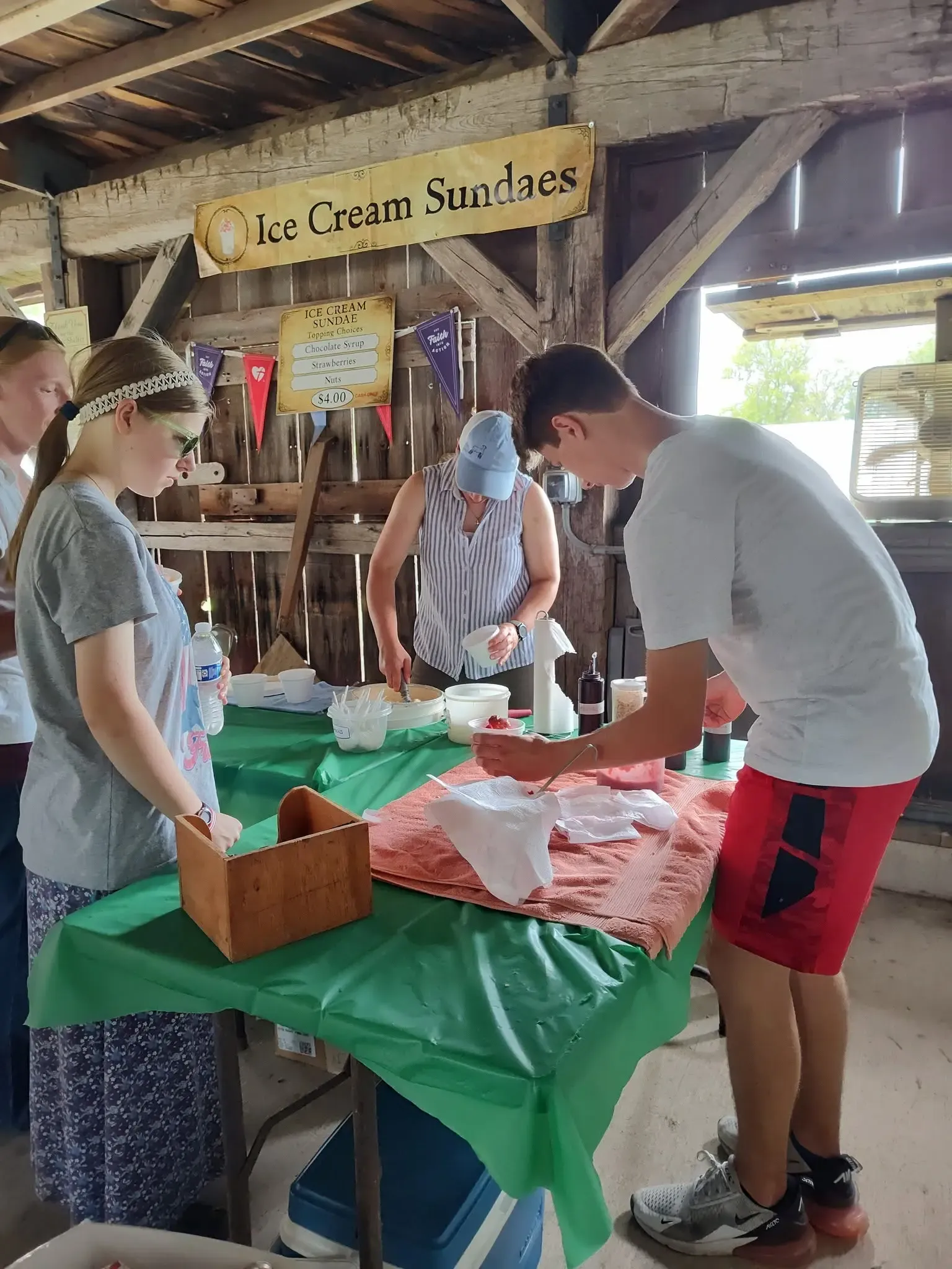 Volunteers scooping ice cream sundaes at the stand beneath the “Ice Cream Sundaes” banner in the barn