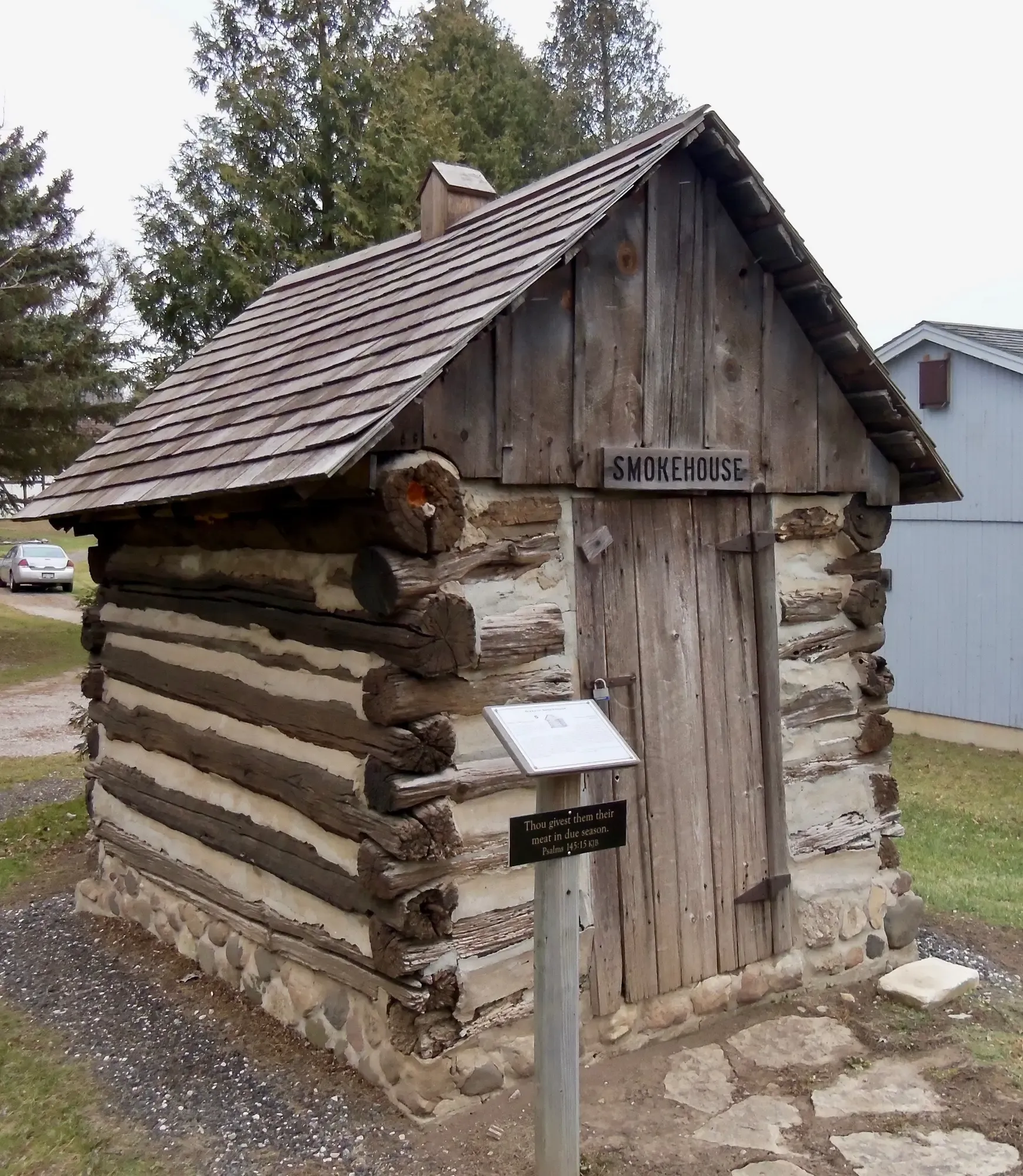 The little log Smokehouse under its cedar-shingled roof and rooftop smoke vent, an interpretive sign on a post by the plank door