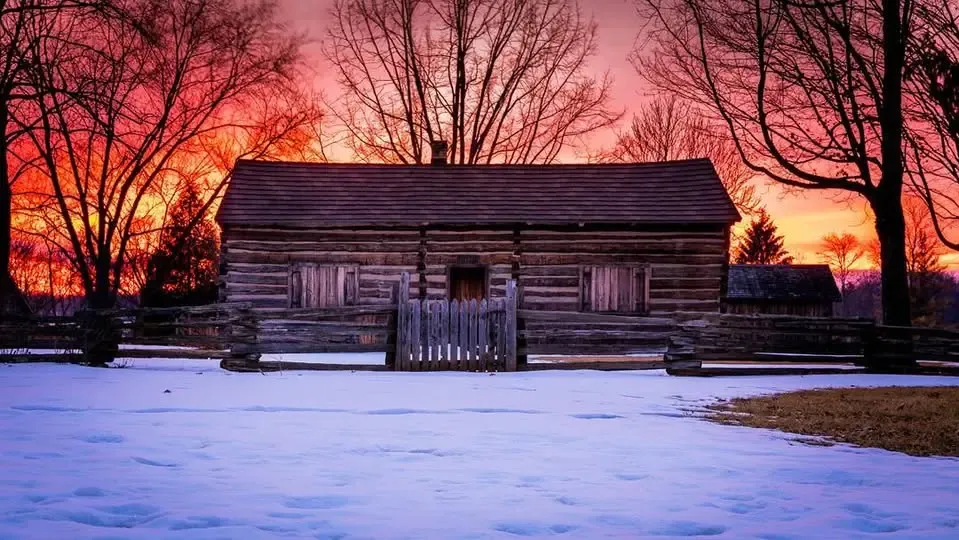The log cabin under snow at sunset, the sky glowing red behind bare trees