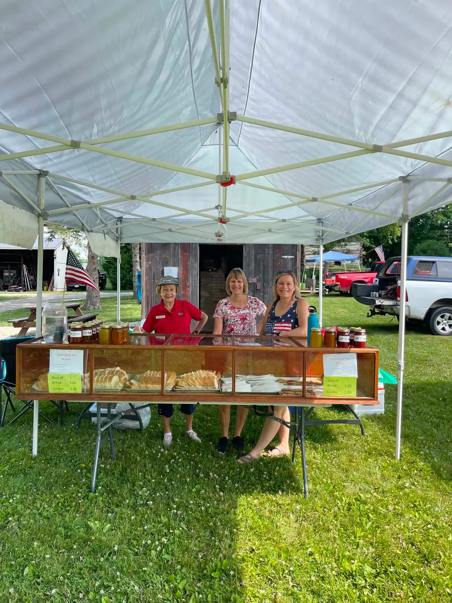 Three volunteers at the bake-sale stand, the vintage glass case filled with sliced bread and lined with jars of homemade jam