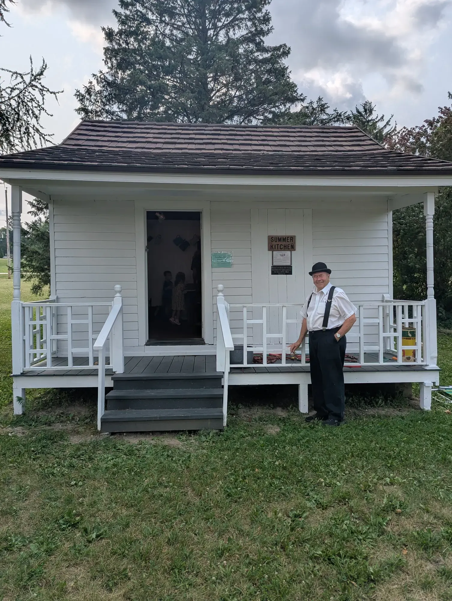 The white clapboard Summer Kitchen with its front porch and "Summer Kitchen" sign, a docent in a period bowler hat and suspenders standing at the porch rail