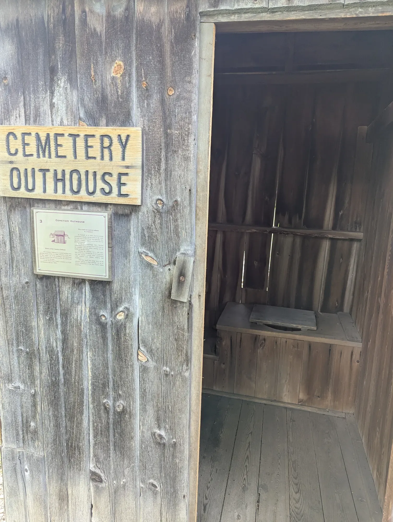 The Cemetery Outhouse with its carved sign and history plaque, the door open on the wooden bench seat inside