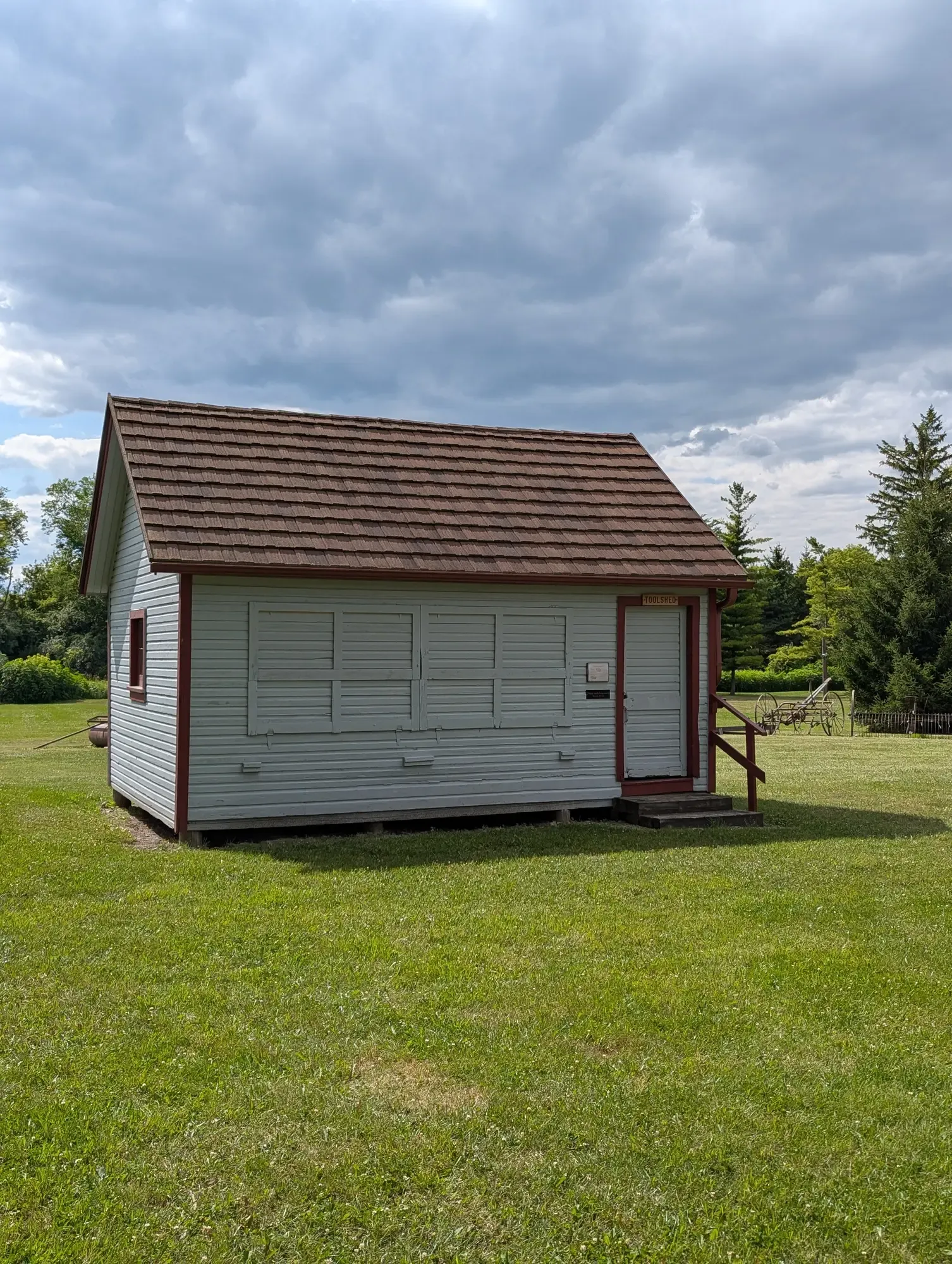 The white clapboard Tool Shed with red trim and a "Toolshed" sign over the door, an antique hay rake on the lawn behind it