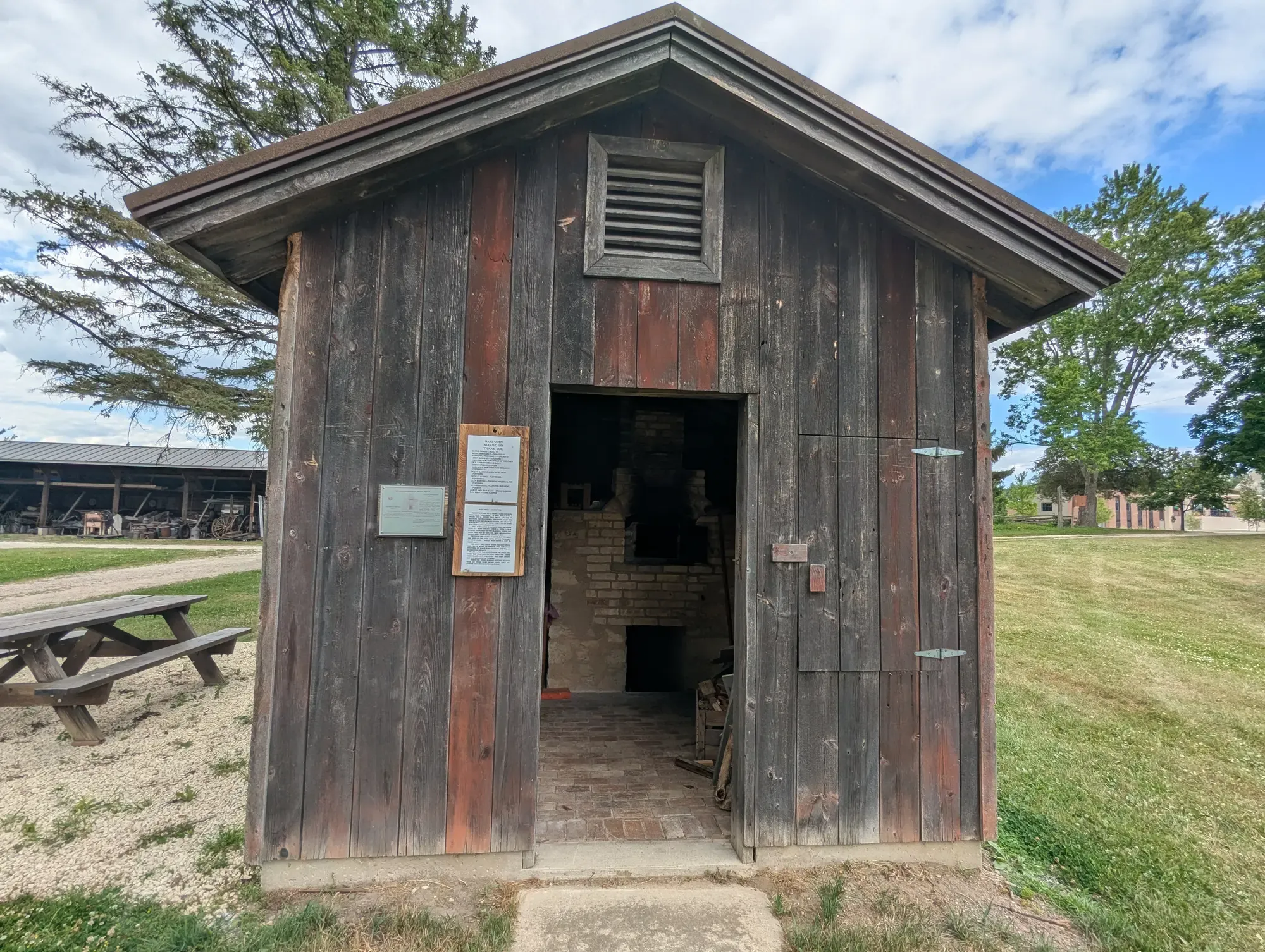 The weathered wooden Bake Oven building with its door open, the brick bake oven visible inside, a picnic table on the gravel beside it