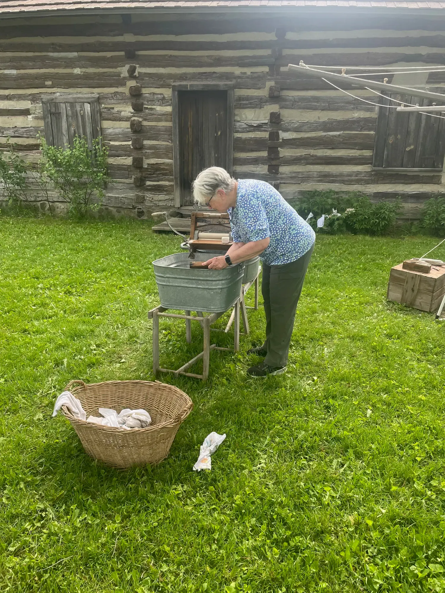 A volunteer feeding wash through a hand-crank wringer behind the log cabin, a wicker laundry basket in the grass